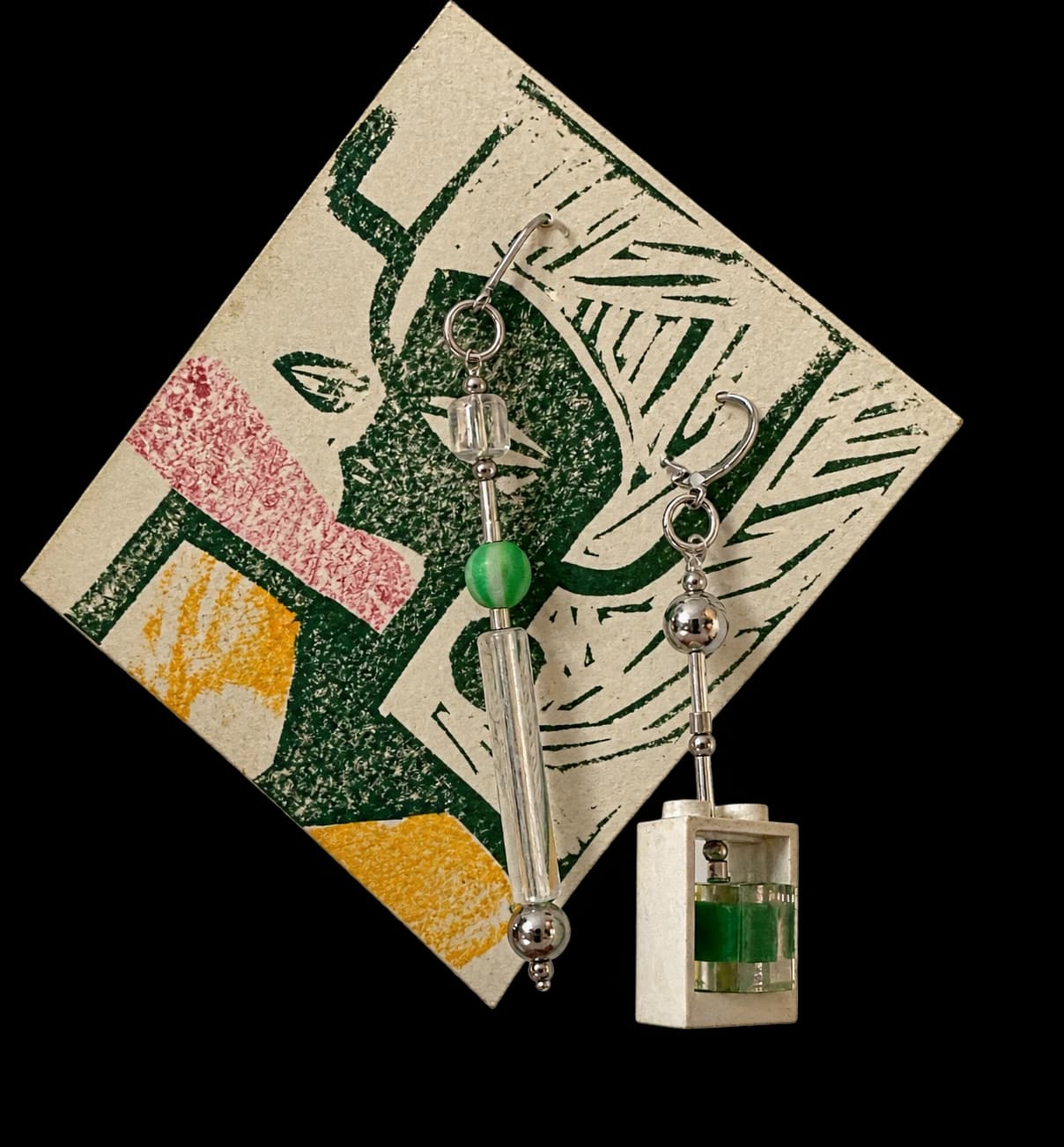 A pair of mismatched handmade earrings, one of glass beads and steel balls, one with a white building brick holding a green bead, hung on a linocut print card