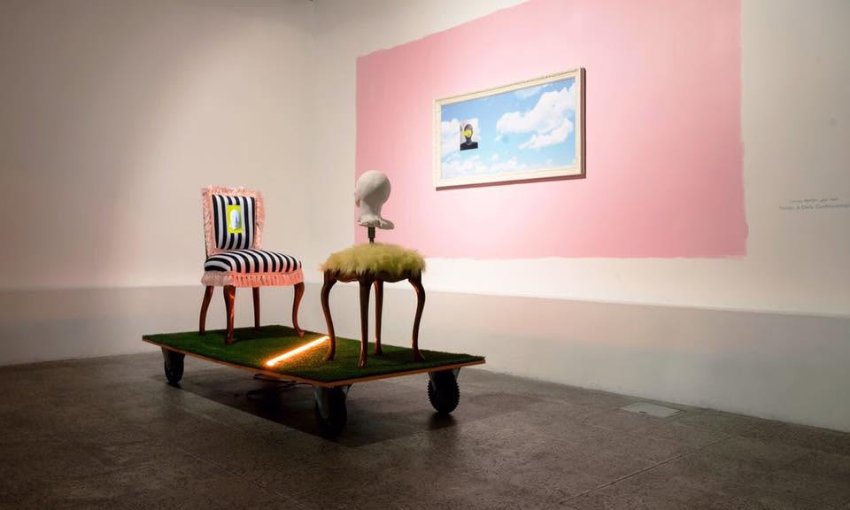 The finished installation in a gallery: a black-and-white striped chair with pink fringe and a mannequin head on a furry stool, on a grass-covered platform on wheels, in front of a pink wall with a framed sky painting.